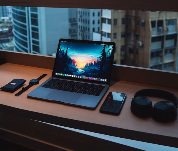 A laptop, phone and headphones set up on a desk by a window